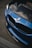 A close-up view of the front part of a blue BMW car, showcasing the iconic BMW emblem and grille. The hood appears to have a carbon fiber finish, and there are reflections visible on the surface. The logo of KZ Auto Group is visible on the bottom part of the bumper.