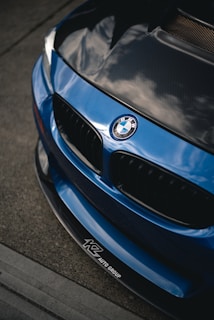 A close-up view of the front part of a blue BMW car, showcasing the iconic BMW emblem and grille. The hood appears to have a carbon fiber finish, and there are reflections visible on the surface. The logo of KZ Auto Group is visible on the bottom part of the bumper.
