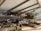 Inside a museum hangar, a vintage biplane with wooden pontoons is displayed prominently. The aircraft is surrounded by sandbags and other historical artifacts, and various other planes are visible in the background. The ceiling and structural beams of the hangar create a spacious atmosphere.