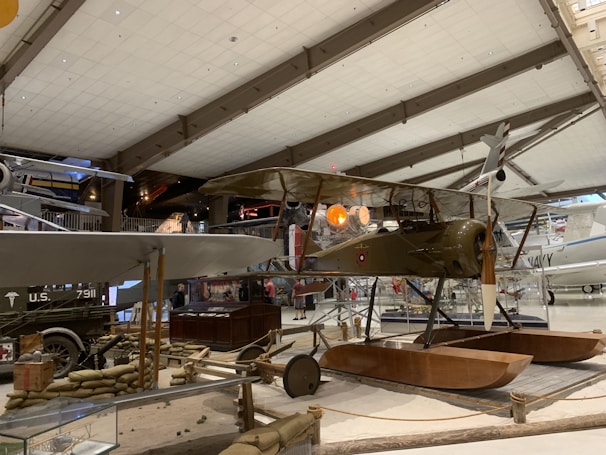 Inside a museum hangar, a vintage biplane with wooden pontoons is displayed prominently. The aircraft is surrounded by sandbags and other historical artifacts, and various other planes are visible in the background. The ceiling and structural beams of the hangar create a spacious atmosphere.