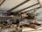 Inside a museum hangar, a vintage biplane with wooden pontoons is displayed prominently. The aircraft is surrounded by sandbags and other historical artifacts, and various other planes are visible in the background. The ceiling and structural beams of the hangar create a spacious atmosphere.