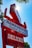 A red and white retro-style sign displays advertisements for hamburgers, emphasizing 'Instant Service' and 'Dick's Deluxe 1/4 lb. 100% Beef.' The sign is set against a clear blue sky with sunlight creating a starburst effect.