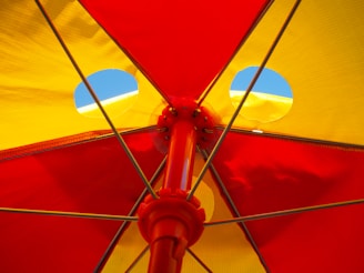 Brightly colored sections of a beach umbrella in red and yellow with metal support rods extending from the center. The sky is visible through circular openings in the umbrella fabric.