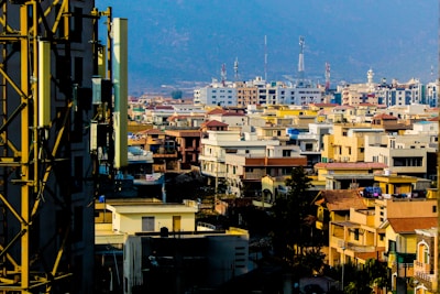 A cityscape featuring a densely packed area of buildings with a variety of architectural styles, including apartment complexes and residential houses. Visible communication towers extend into the sky, and the backdrop consists of hazy mountains.