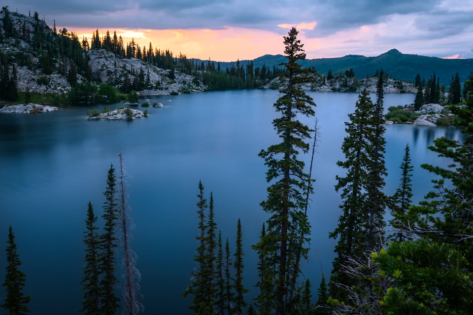Serene lake nestled in forested area, surrounded by trees reflecting sunset's warm glow