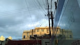 A building with multiple windows is topped with various antennas and satellite dishes. Numerous electrical wires crisscross in the foreground, connected to a utility pole. The building is partially reflected in a large glass surface on the right. The sky above is cloudy, with patches of sunlight illuminating parts of the structure.