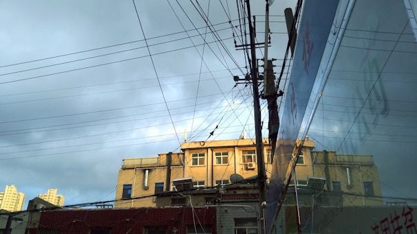 A building with multiple windows is topped with various antennas and satellite dishes. Numerous electrical wires crisscross in the foreground, connected to a utility pole. The building is partially reflected in a large glass surface on the right. The sky above is cloudy, with patches of sunlight illuminating parts of the structure.