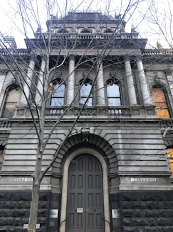An old, grand university building with large columns and arched windows. The architecture is adorned with intricate stonework and a prominent entrance under bare tree branches.