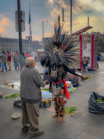 A person dressed in traditional Aztec attire with an elaborate feathered headdress, a symbolic skull, and clothing featuring intricate patterns is interacting with another individual. Surrounding them are vendors and visitors, contributing to a lively cultural scene. In the background, a large flag and historical architecture are visible under a partly cloudy sky.