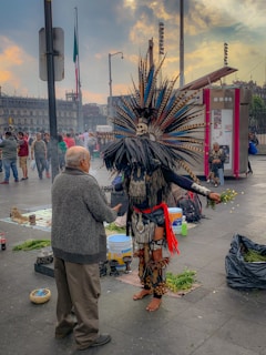 A person dressed in traditional Aztec attire with an elaborate feathered headdress, a symbolic skull, and clothing featuring intricate patterns is interacting with another individual. Surrounding them are vendors and visitors, contributing to a lively cultural scene. In the background, a large flag and historical architecture are visible under a partly cloudy sky.
