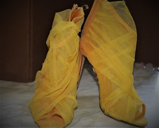 A pair of high-heeled shoes covered with wrapped yellow gauze or mesh. The shoes have an open-toe design and are set against a muted background, possibly a floor. The wrapping gives them a unique and artistic appearance with a textured surface.