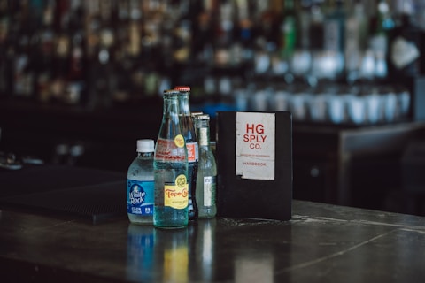 Several bottles and a menu or notebook are arranged on a bar counter. The bottles include a club soda and a Topo Chico mineral water, and they are positioned next to a dark-covered book labeled 'HG Sply Co. Handbook'. The background is blurred, showing numerous other bottles on shelves, typical of a bar setting.