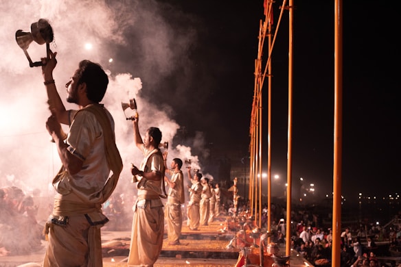 Several people dressed in traditional attire perform a ritual at night. They hold metal vessels emitting smoke. The scene is illuminated by artificial lights in the background, while a crowd observes the performance.
