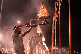 A group of individuals are performing a traditional ritual involving large, tiered brass lamps. They are dressed in similar attire and are standing in a line, holding the lamps high as they emit a warm glow and smoke. The background is dark, accentuating the bright flames and creating a mystical atmosphere.