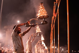 A group of individuals are performing a traditional ritual involving large, tiered brass lamps. They are dressed in similar attire and are standing in a line, holding the lamps high as they emit a warm glow and smoke. The background is dark, accentuating the bright flames and creating a mystical atmosphere.