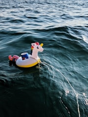 A colorful unicorn float carrying a can is floating on the water with gentle ripples. The float has a rainbow mane and tail, with a yellow body. The background features expansive, shimmering water under soft lighting, creating a serene setting.