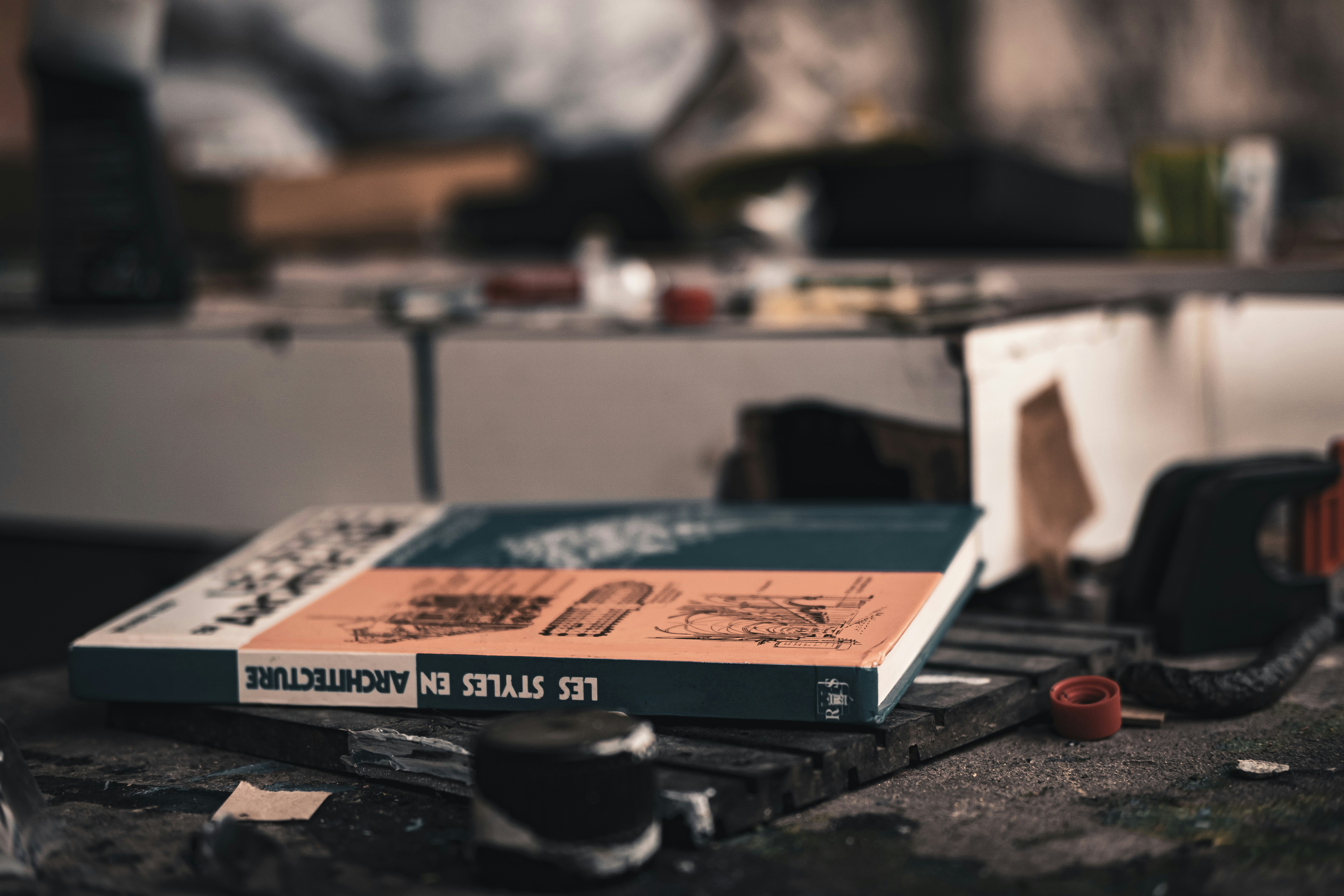A book titled 'Les Styles en Architecture' rests on a cluttered tabletop, surrounded by various objects and papers. The book's cover features architectural sketches highlighted in orange and white against a teal background. The ambiance suggests a creative or work-in-progress environment.