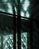A modern architectural interior featuring a ceiling with patterned tiles and geometric grids. The space is accentuated by dramatic lighting and shadow play on the walls and ceiling.