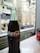 A classic glass bottle of Coca-Cola is placed on a smooth, reflective surface with a blurred cityscape visible through a window in the background. To the right, a partial view of a Coca-Cola can features its signature red label.