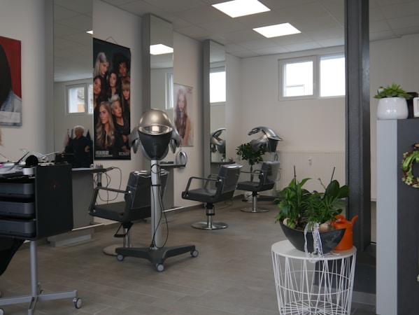 A modern hair salon interior with styling chairs, hair drying equipment, and large posters featuring hairstyles on the walls. There are potted plants adding a touch of greenery to the sleek and clean design. The room is well-lit with natural light coming through the windows and artificial ceiling lights.