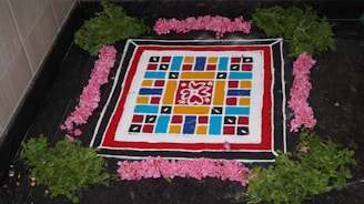 A colorful, symmetrical rangoli design is created on a dark surface, consisting of geometric patterns and vibrant colors. The design features a red, white, and black border, with multicolored squares and an intricate floral motif in the center. Surrounding the design are freshly picked green leaves and pink flowers arranged neatly in a square pattern.