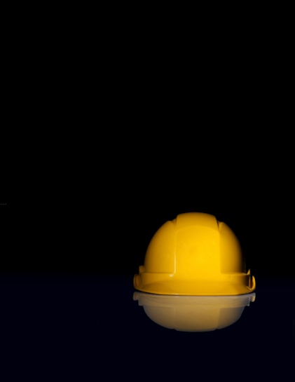 A bright yellow hard hat is centrally placed against a stark black background, creating a striking contrast. The hat's glossy surface reflects some light, enhancing its visibility. Its reflection can be seen subtly below.