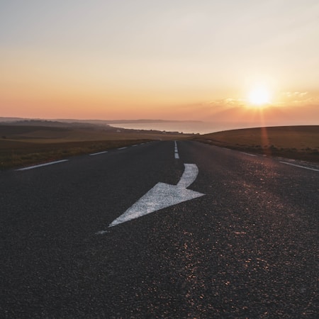 A straight road stretches into the distance with a large white arrow painted on the asphalt, directing to the right. The road is flanked by open fields. The sky is filled with the warm hues of a sunset, casting a serene glow over the landscape.