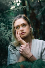 A person with blonde hair and contemplative expression is surrounded by lush green ferns in a dense, forested environment. The soft lighting and natural setting create an intimate and serene atmosphere.