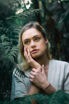 A person with blonde hair and contemplative expression is surrounded by lush green ferns in a dense, forested environment. The soft lighting and natural setting create an intimate and serene atmosphere.