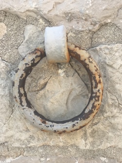 An old, weathered metal ring anchored into a rough stone wall. The metal shows significant rust and wear, contributing to an overall aged appearance. The stone wall is textured with cracks and various shades of gray and beige.