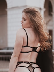 A young woman with long, wavy hair is seen from the back. She is wearing black lingerie with sheer, polka dot details. The background is urban, featuring an archway and a railing with a blurred car and building.