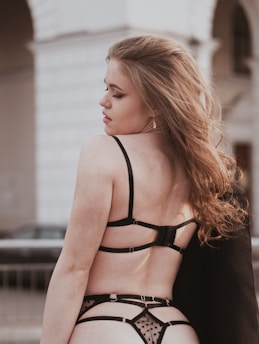 A young woman with long, wavy hair is seen from the back. She is wearing black lingerie with sheer, polka dot details. The background is urban, featuring an archway and a railing with a blurred car and building.