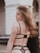 A young woman with long, wavy hair is seen from the back. She is wearing black lingerie with sheer, polka dot details. The background is urban, featuring an archway and a railing with a blurred car and building.