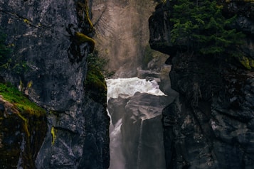 A powerful waterfall cascades between two towering rocky cliffs, surrounded by lush greenery. Mist rises from the base of the falls, creating a mystical atmosphere as the water forcefully travels down. The moss-covered rocks and thick trees add a sense of wilderness and untouched nature.