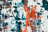 Abstract painting showcasing a blend of bright and vibrant colors with a predominantly chaotic texture. The colors include splashes of teal, orange, white, and intermittent darker and lighter shades spread across the canvas. Thick, overlapping brushstrokes create a textural depth and an intricate pattern.