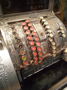 A vintage cash register with ornate metallic designs and multiple rows of circular keys featuring various monetary values in old currency units like pounds, shillings, and pence. The keys are predominantly black, red, and cream-colored, and the device appears to be a classic example of early 20th-century craftsmanship.