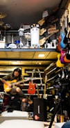 A musician seated, engrossed in playing an electric guitar amidst a vibrant environment filled with musical instruments. Various guitars in diverse colors hang on the wall, and keyboards are neatly stacked. Above, there is a display of percussion instruments including drums and other equipment.