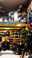 A musician seated, engrossed in playing an electric guitar amidst a vibrant environment filled with musical instruments. Various guitars in diverse colors hang on the wall, and keyboards are neatly stacked. Above, there is a display of percussion instruments including drums and other equipment.