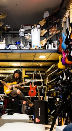 A musician seated, engrossed in playing an electric guitar amidst a vibrant environment filled with musical instruments. Various guitars in diverse colors hang on the wall, and keyboards are neatly stacked. Above, there is a display of percussion instruments including drums and other equipment.