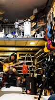 A musician seated, engrossed in playing an electric guitar amidst a vibrant environment filled with musical instruments. Various guitars in diverse colors hang on the wall, and keyboards are neatly stacked. Above, there is a display of percussion instruments including drums and other equipment.