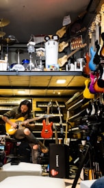 A musician seated, engrossed in playing an electric guitar amidst a vibrant environment filled with musical instruments. Various guitars in diverse colors hang on the wall, and keyboards are neatly stacked. Above, there is a display of percussion instruments including drums and other equipment.