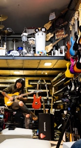 A musician seated, engrossed in playing an electric guitar amidst a vibrant environment filled with musical instruments. Various guitars in diverse colors hang on the wall, and keyboards are neatly stacked. Above, there is a display of percussion instruments including drums and other equipment.