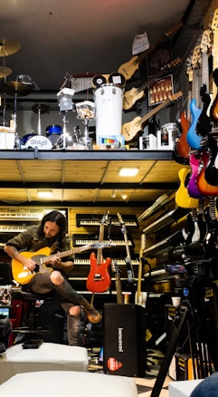 A musician seated, engrossed in playing an electric guitar amidst a vibrant environment filled with musical instruments. Various guitars in diverse colors hang on the wall, and keyboards are neatly stacked. Above, there is a display of percussion instruments including drums and other equipment.