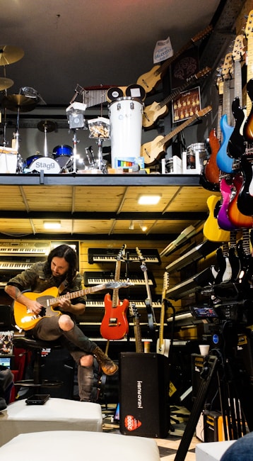 A musician seated, engrossed in playing an electric guitar amidst a vibrant environment filled with musical instruments. Various guitars in diverse colors hang on the wall, and keyboards are neatly stacked. Above, there is a display of percussion instruments including drums and other equipment.