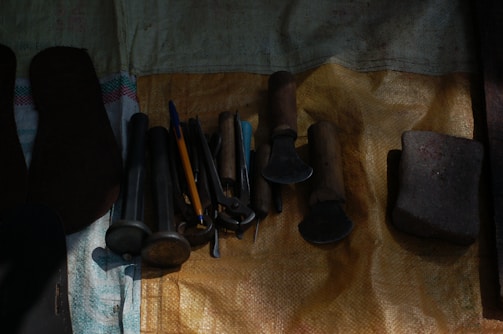 A collection of various tools laid out on a textured surface. The tools appear to include hammers, files, and chisels with wooden handles, alongside a pen and a stone object. The surface is covered with a woven material of varying shades.
