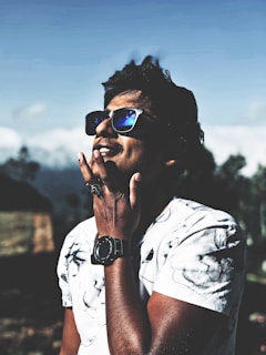 A person wearing reflective sunglasses and a patterned white t-shirt is enjoying the outdoors, exuding a sense of relaxation and style. The individual has short, dark hair and is thoughtfully touching their chin while standing against a blurred natural background.