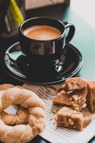 A black cup of coffee sits on a matching saucer alongside pastries. The background features text on a piece of paper, partially obscured by a broken crumbly dessert and a twisted bread roll.