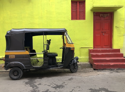 An auto-rickshaw is parked by a vibrant lime green building with a red door and steps. The auto-rickshaw is black and yellow, contrasting with the vivid colors of the building.