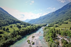 The image features a scenic landscape with a winding river cutting through a lush valley surrounded by green hills and mountains. The clear blue sky is dotted with a few fluffy clouds, and the sunlight casts vibrant shadows on the verdant fields and trees lining the riverbank.