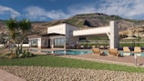 A modern house is situated in a scenic mountainous backdrop. The structure is a single-story building with large windows and stone accents. In the foreground, there is a swimming pool surrounded by a patio with wooden lounge chairs. The landscape features well-manicured grass, desert plants, and a section of decorative gravel.
